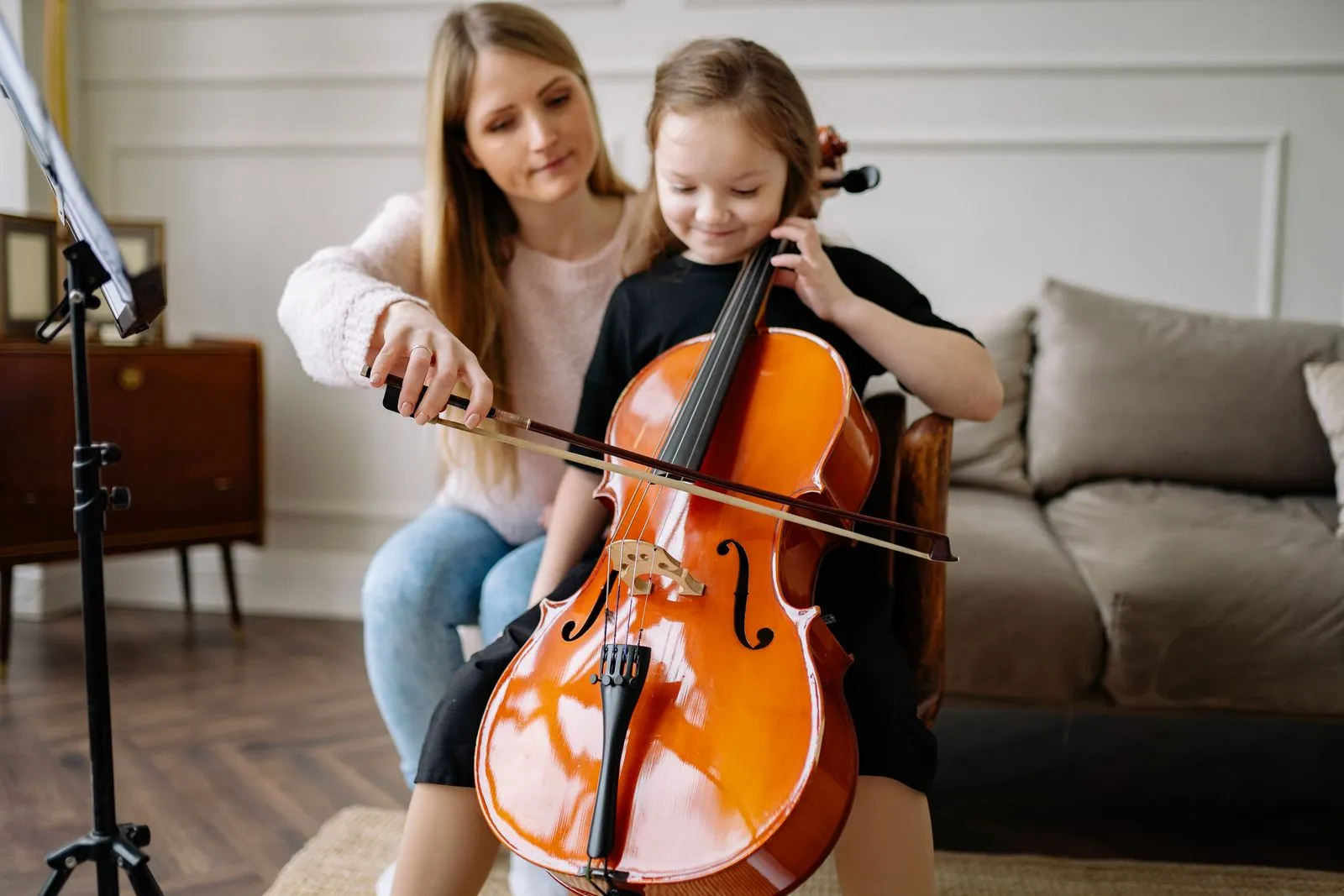 A woman guides her daughter in playing the cello, creating a musical learning experience indoors.